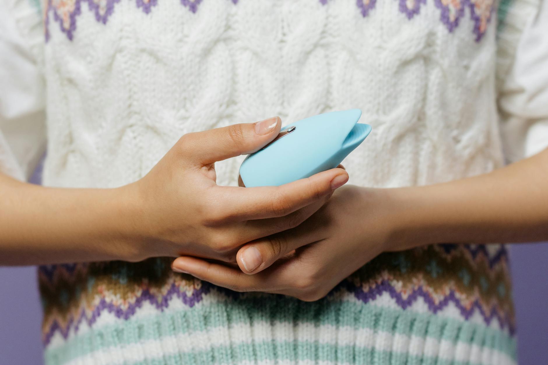 Mãos segurando um vibrador azul contra um suéter de tricô, representando toque compartilhado e intimidade